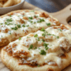 A rustic Creamy Chicken Alfredo Pizza sits on a light maple wooden board. The golden crust is crisp at the edges, topped with a glossy white Alfredo sauce, shredded chicken, wilted spinach, and melted mozzarella. A light sprinkle of parmesan and a basil leaf add color, while a subtle drizzle of olive oil and pepper flakes hint at depth. The scene is warm, inviting, and homey with soft shadows and natural kitchen lighting.