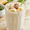 A frothy pale yellow banana milk in a tall clear glass on a light wooden table, sliced banana and a drizzle of condensed milk on the side, a pinch of cinnamon, a mint leaf for color, and a small glass of water in the background; clean, comforting, modern presentation.