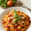 A bright plate of creamy tomato pasta with glossy tomato cream sauce, ribbons of al dente pasta, grated parmesan, fresh basil, and a drizzle of olive oil. The dish is plated on a white round plate on a light wooden table, with a few basil leaves, chili flakes, and parmesan shavings nearby, creating a rustic yet modern presentation.