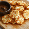 Golden brown crispy cottage cheese chips arranged on a white plate with a sprinkle of paprika and chopped herbs, set on a light wooden board with a small dish of dip in the background; airy, inviting, and savory with cheese flecks visible on the surface.