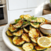 Golden-brown crispy zucchini chips fanned on a white plate, their edges slightly curled and crackly. Thin, uniformly sliced green spears coated with a light parmesan-crumb crust and a whisper of sea salt. A small bowl of tangy salt-and-vinegar dip, lemon zest, and dill garnish complete a vibrant, fresh-snack presentation on a rustic wooden board.