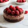 A decadent dark chocolate raspberry mousse cake on a white plate, glossy ganache topping, fresh raspberries, and a dusting of cocoa. Layered slices reveal a dark chocolate cake base and a silky raspberry mousse, styled on a neutral placemat with subtle speckled surface.
