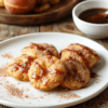 Golden brown apple fritter bites on a white plate dusted with cinnamon sugar, small apple pieces visible in a fluffy batter, a light maple glaze drizzle, warm autumn lighting on a rustic wooden board.