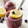Bright blueberry lemon sorbet in a clear glass bowl, vibrant blue-purple hue with lemon zest flecks, surrounded by fresh blueberries and lemon slices on a white marble surface, a mint sprig for garnish, glassy surface with icy crystals, simple rustic plating.