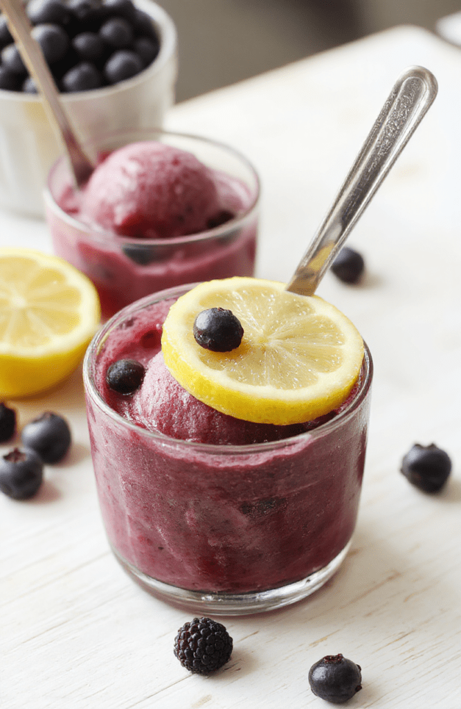 Bright blueberry lemon sorbet in a clear glass bowl, vibrant blue-purple hue with lemon zest flecks, surrounded by fresh blueberries and lemon slices on a white marble surface, a mint sprig for garnish, glassy surface with icy crystals, simple rustic plating.