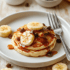 A stack of golden fluffy mini banana pancakes on a white plate with maple syrup drizzle, sliced bananas, fresh berries, a sprig of mint, and a light dusting of cinnamon on a clean wooden table bathed in warm morning light.