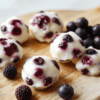 Bright white plate filled with twelve frozen blueberry yogurt bites, each swirled with creamy yogurt and dotted with blueberries. The bites rest on a pale wooden board with a few fresh blueberries and a light mint leaf for garnish. The surface is clean and airy, with soft natural light highlighting the smooth texture and blueberry specks inside the bites.