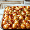Golden bubbly pizza tot casserole in a square baking dish with crispy tots, melted mozzarella, pepperoni circles, and colorful diced peppers. A sprig of basil adds color on top, with marinara visible beneath the tots and a lightly browned crust around the edges; the dish sits on a rustic wooden table with a slice plated to reveal gooey cheese and crispy edges.