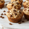 Golden fluffy Greek yogurt muffins with blueberries, stacked on a white plate. A drizzle of honey, a light cinnamon dusting, and oats in the background create a wholesome, breakfast-ready scene on a light wooden table.