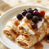 Golden, fluffy oatmeal crepes rolled around fresh berries and yogurt, draped with maple syrup, dusted with cinnamon, served on a light wooden board with a small jar of berries and a fork, bright natural light and soft shadows.