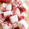 Fluffy pink raspberry marshmallows arranged on a white plate dusted with powdered sugar, fresh raspberries nearby, a light marble backdrop and soft natural light highlight the airy texture and delicate finish.