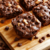 Fudgy chocolate chip cookie brownies plated in a square pan, cut into neat squares. Rich dark brownie crust with glossy surface and pockets of melted chocolate, swirled with chunks of chocolate chips. Golden-brown cookie dough bits peek through. The bars sit on a light wooden board with a dusting of flaky sea salt and a drizzle of extra chocolate. Warm, inviting, and cozy dessert styling with soft natural light.