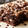 Luxurious fudgy yogurt brownies plated on a white square dish atop a light wood board. Rich dark chocolate crumb with a glossy surface, a dollop of yogurt glaze, cocoa powder dusting, and a mint leaf accent create a simple, inviting dessert scene in natural light.
