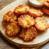 A close-up shot of golden crispy hash browns stacked on a white plate, flecked with black pepper; a bright yolk peeks from a side of eggs, and a sprinkle of chopped parsley adds color. The plate sits on a light wooden table, with warm morning light highlighting the crackly crust and fluffy interior.