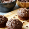 Six fluffy chocolate muffins with glossy tops sit on a light wooden board, cocoa dusting on the surface, a small bowl of yogurt and a few berries in the background, warm natural lighting and rustic plating.