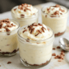 Layered no-bake cheesecake jars with a creamy pale filling, a crumbly graham crust at the bottom, and a glossy berry topping. Served in clear glass jars on a light wooden board, the scene is bright and fresh with soft natural light and a clean, minimalist styling.