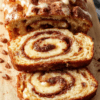 A tall loaf of Moist Cinnamon Roll Banana Bread sits on a rustic wood board, sliced to reveal swirls of cinnamon and creamy glaze. Warm spices dust the crust, a pat of butter melts on a slice, and a cup of coffee adds cozy contrast.