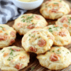 Golden-brown savory ham and cheese cookies on a slate board, flaky butter dough with diced ham and melted cheddar, light herbs, served warm with a small bowl of mustard; rustic wooden table background with soft daylight.