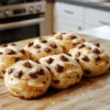 Three golden-brown, pillowy soft milk buns resting on a parchment-lined baking sheet, each studded with melty chocolate chips on top, dusted lightly with powdered sugar, on a rustic wooden board,自然 light, shallow depth of field, bottom third empty for text overlay.