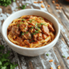 A white bowl holds glossy beef noodles tossed in a dark, spicy sauce, with tender beef strips, bright yellow noodles, red chili slices, and chopped green onions. Sesame seeds speckle the top, cilantro leaves add a pop of green, all arranged on a warm wooden table with a light backdrop.