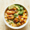 A colorful bowl featuring charred chicken pieces, street corn kernels tossed with lime and cotija, fluffy cilantro-lime rice, avocado slices, black beans, and pico de gallo, finished with fresh cilantro and a lime wedge on a rustic wooden table.